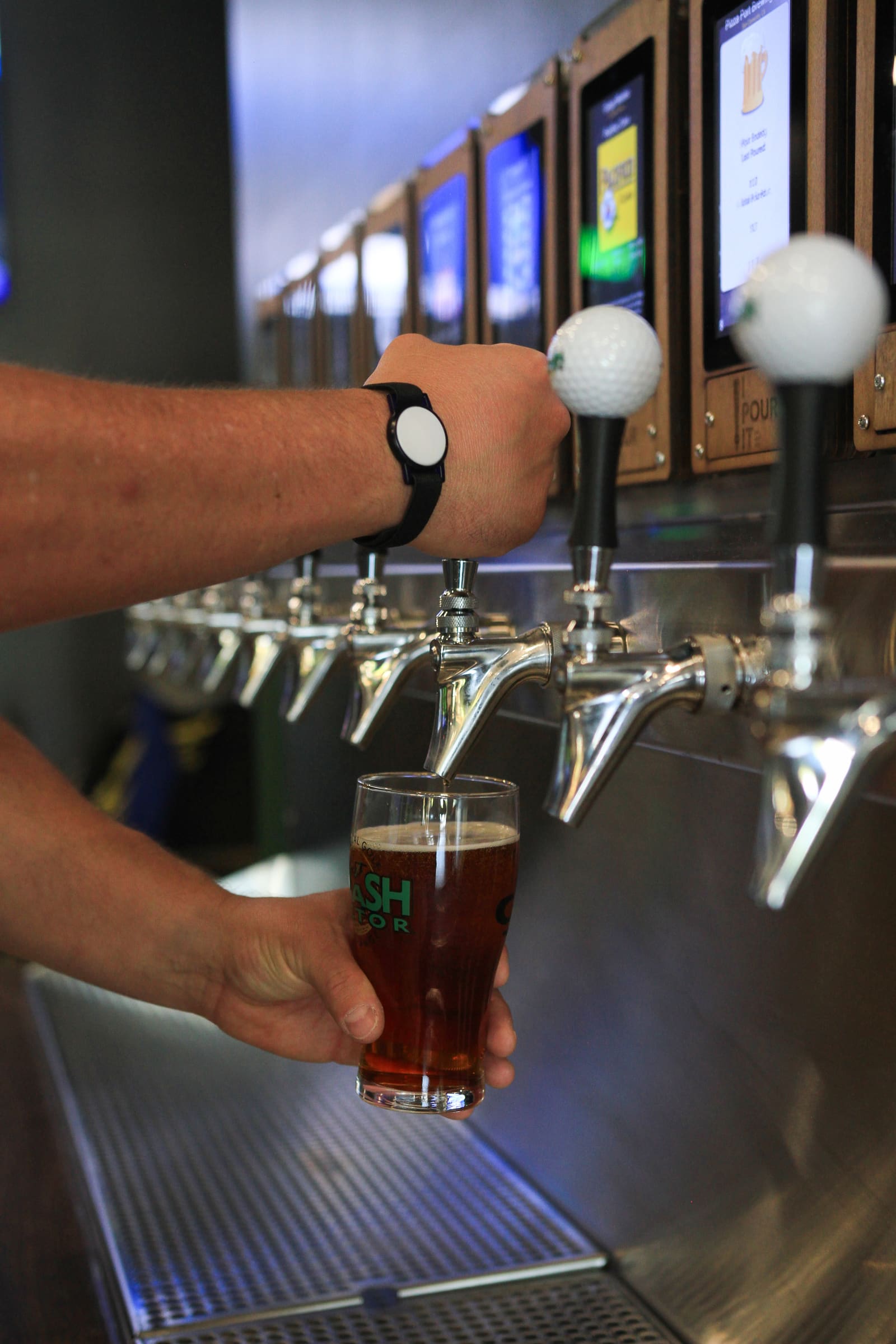 A hand wearing the RFID wristband pouring a beer into a Smash Factor glass, the golf-ball tap handles in a row above.